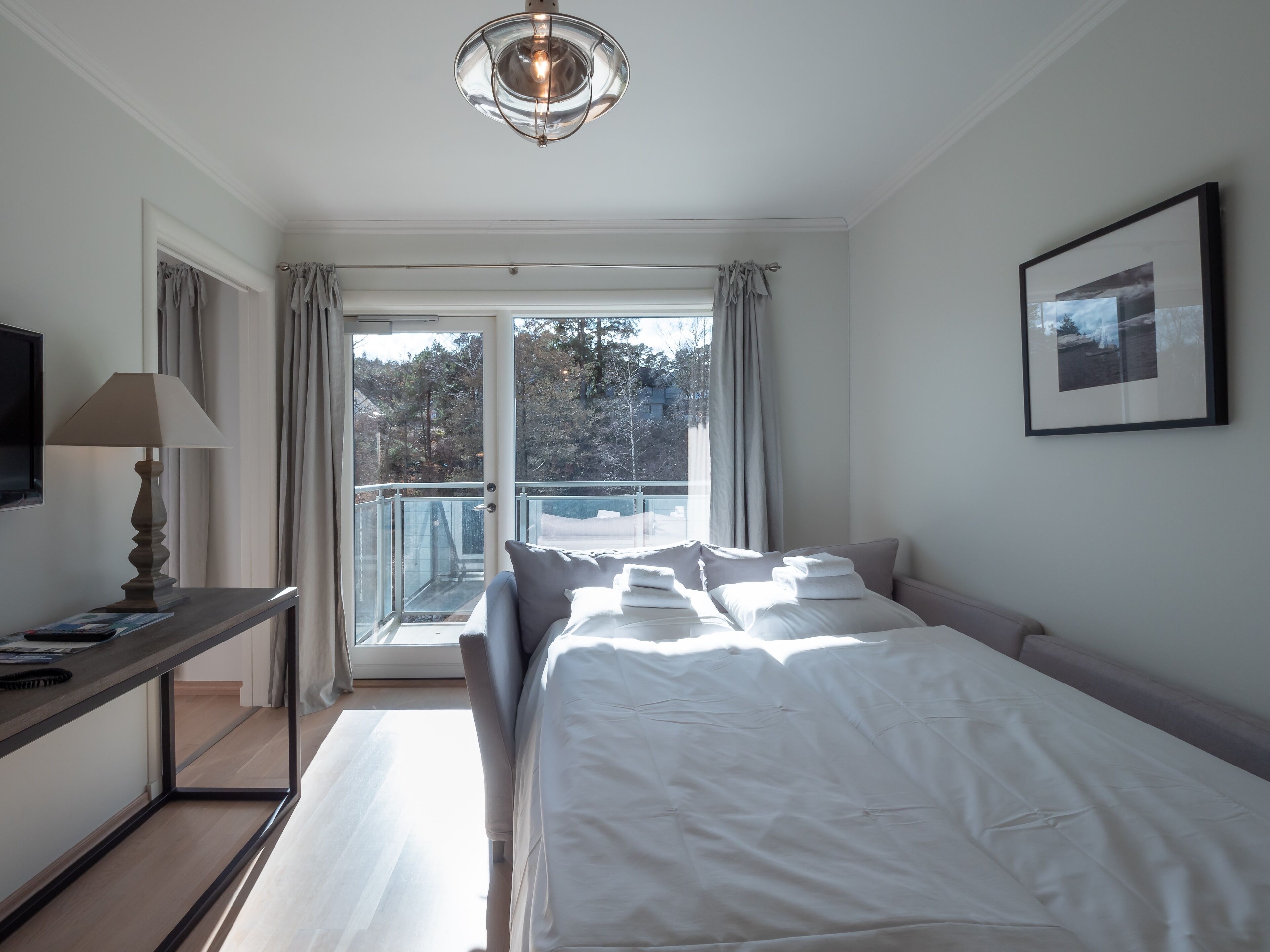 A hotel room with a bed, a desk with a lamp, a television, and a balcony with curtains.