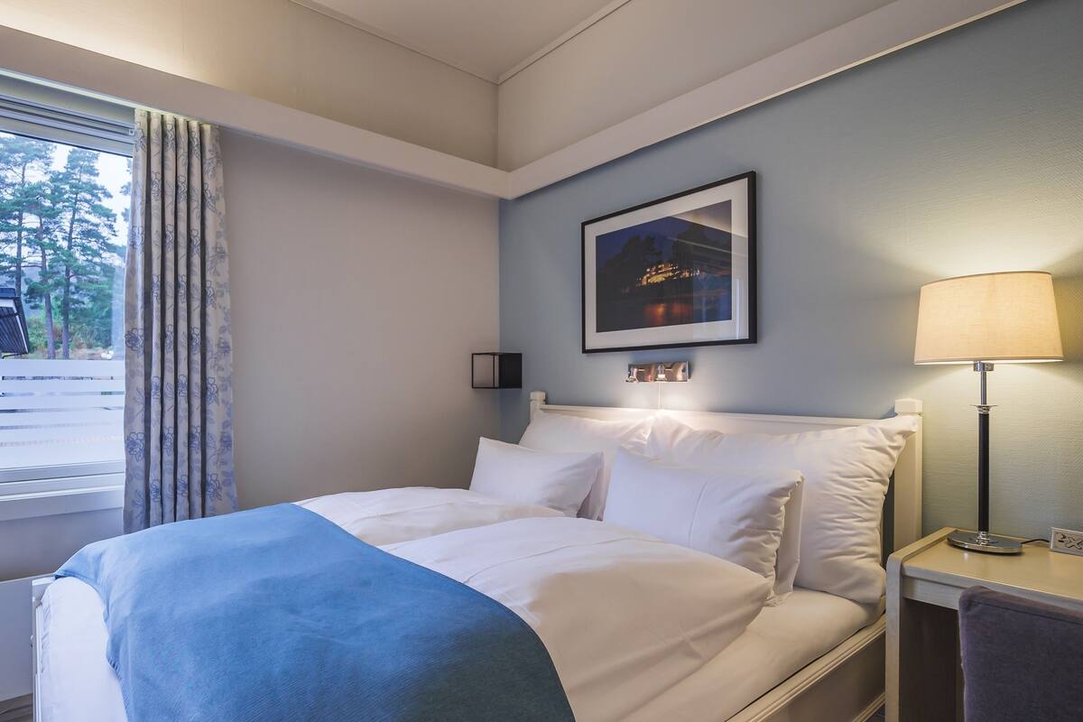 Una cama bien hecha con ropa de cama blanca y azul, un cuadro enmarcado en la pared, una lámpara de noche y una ventana con cortinas.