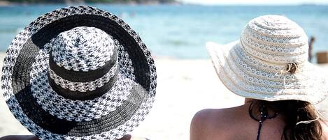 Two people from behind, wearing large, patterned hats, standing on a beach with a view of the sea and a distant shoreline.