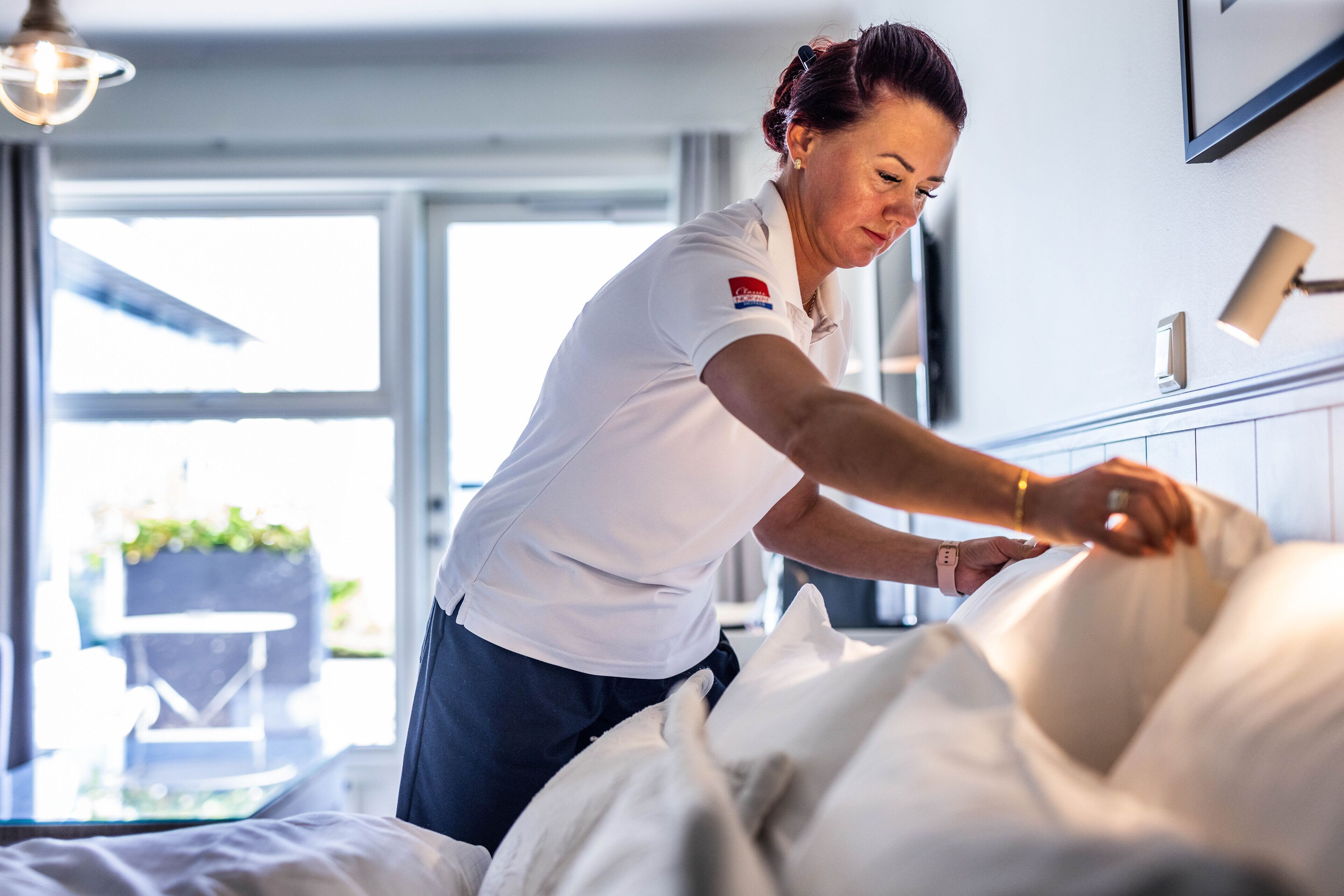 A person adjusting a bed in a room with a window, a chair, and a plant visible in the background.
