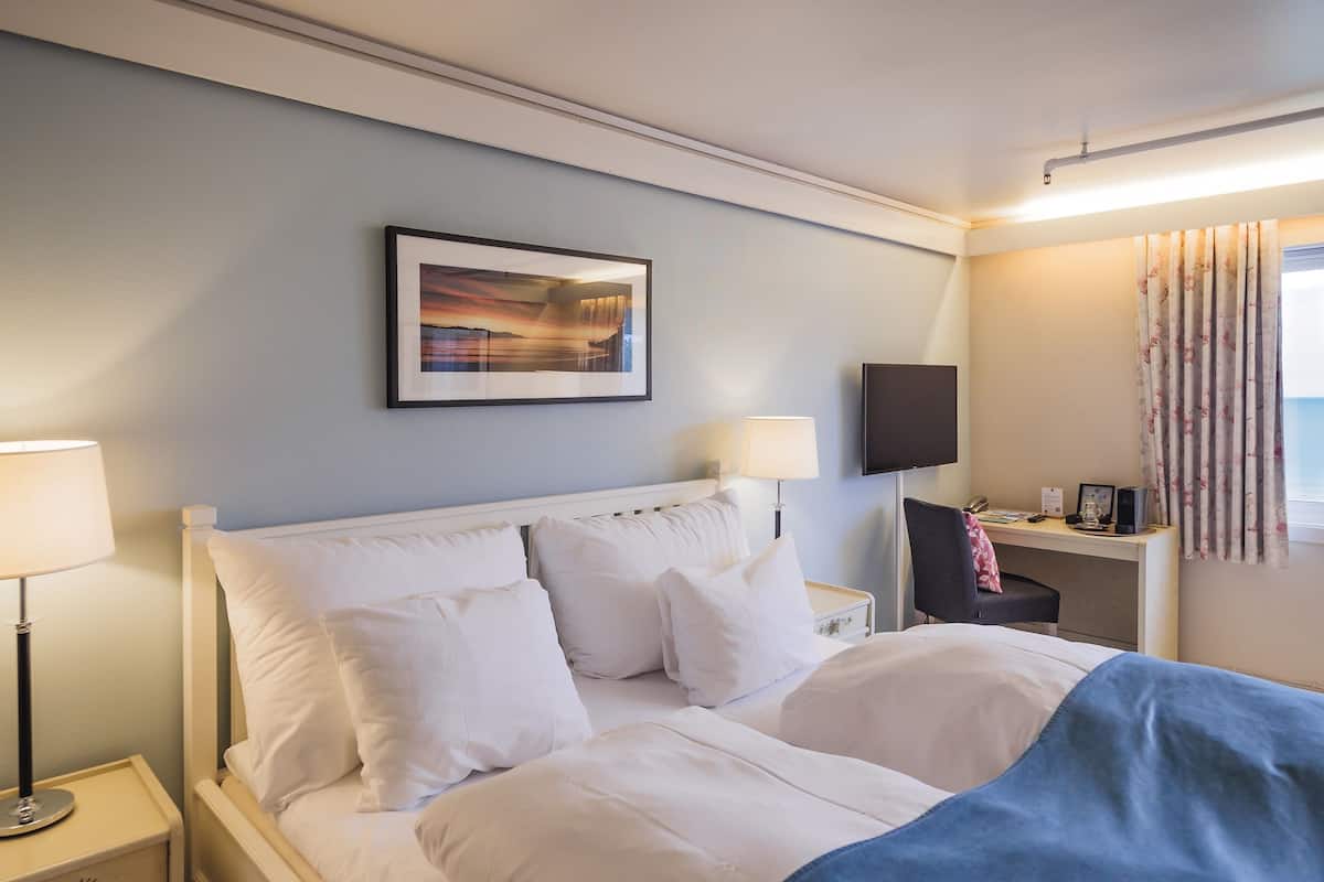 Una cama bien hecha con sábanas blancas, una mesita de noche con lámpara, un cuadro enmarcado en la pared, un escritorio con silla y una ventana con cortinas.