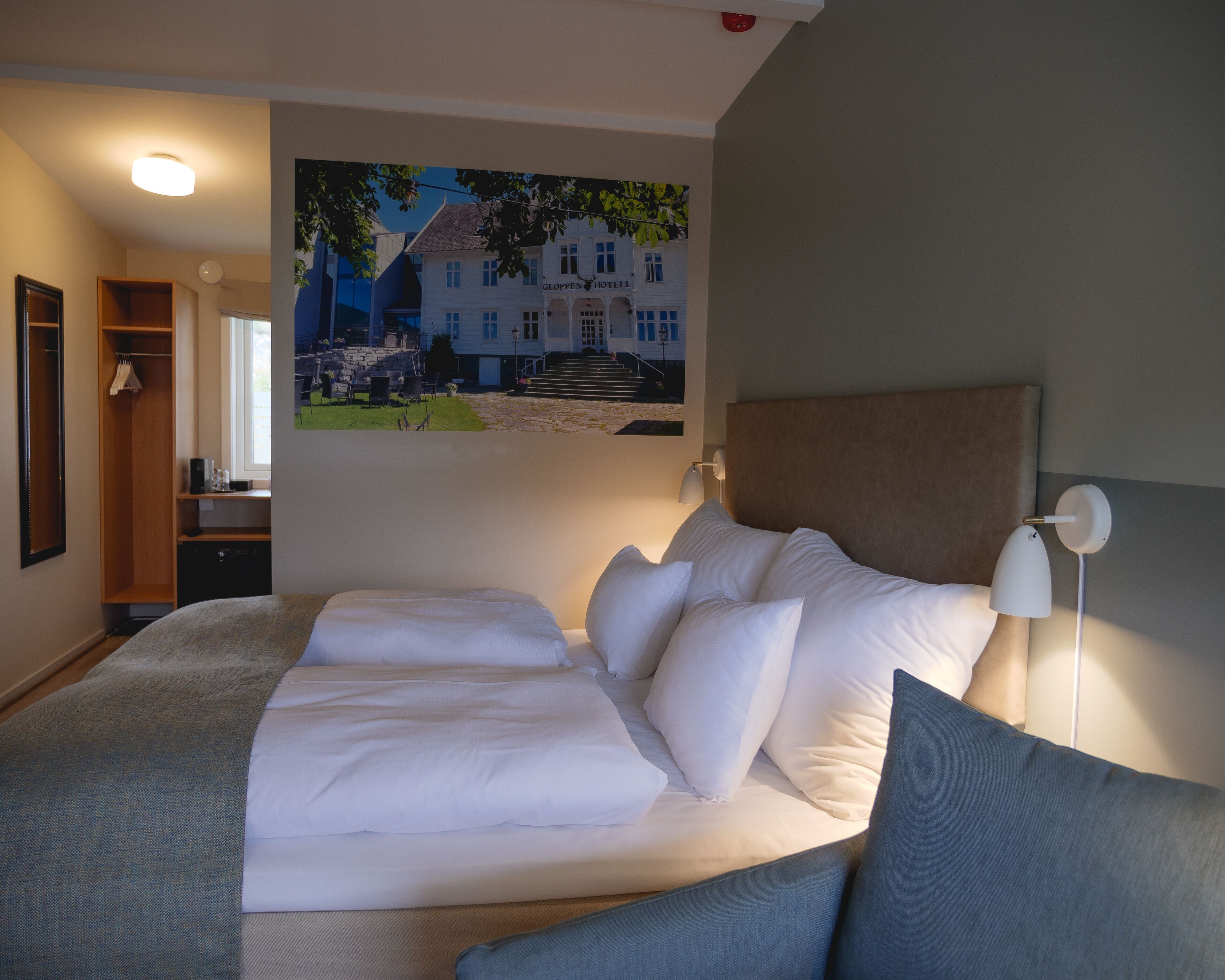 Una habitación de hotel con una cama grande, cabecera, mesita de noche, lámpara y un cuadro de un edificio.
