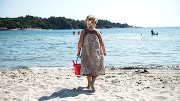 Escena de playa con una persona vestida con un vestido estampado caminando sobre la arena, sosteniendo un cubo rojo.