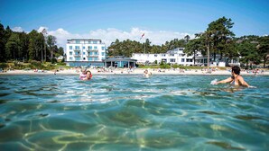 Escena de platja amb aigua cristal·lina, gent nedant i edificis al fons.