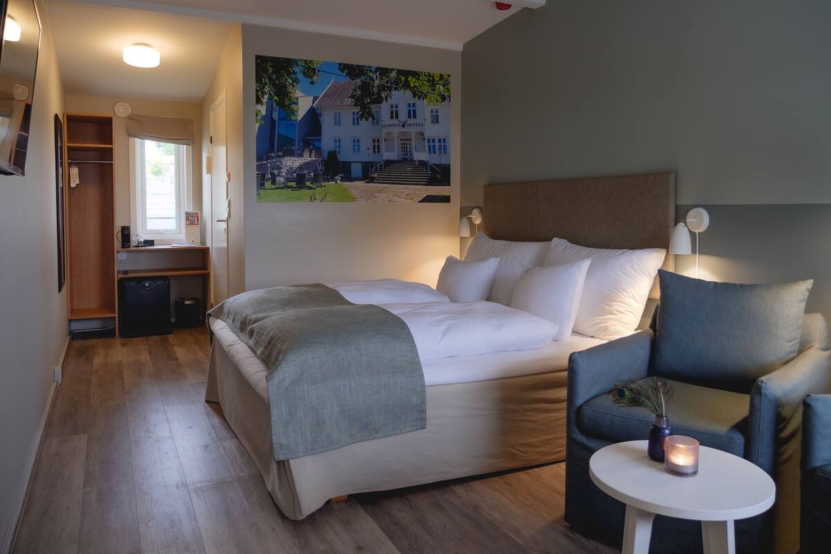 Habitación de hotel con cama, una silla, una mesita con una vela, un cuadro en la pared y un armario.