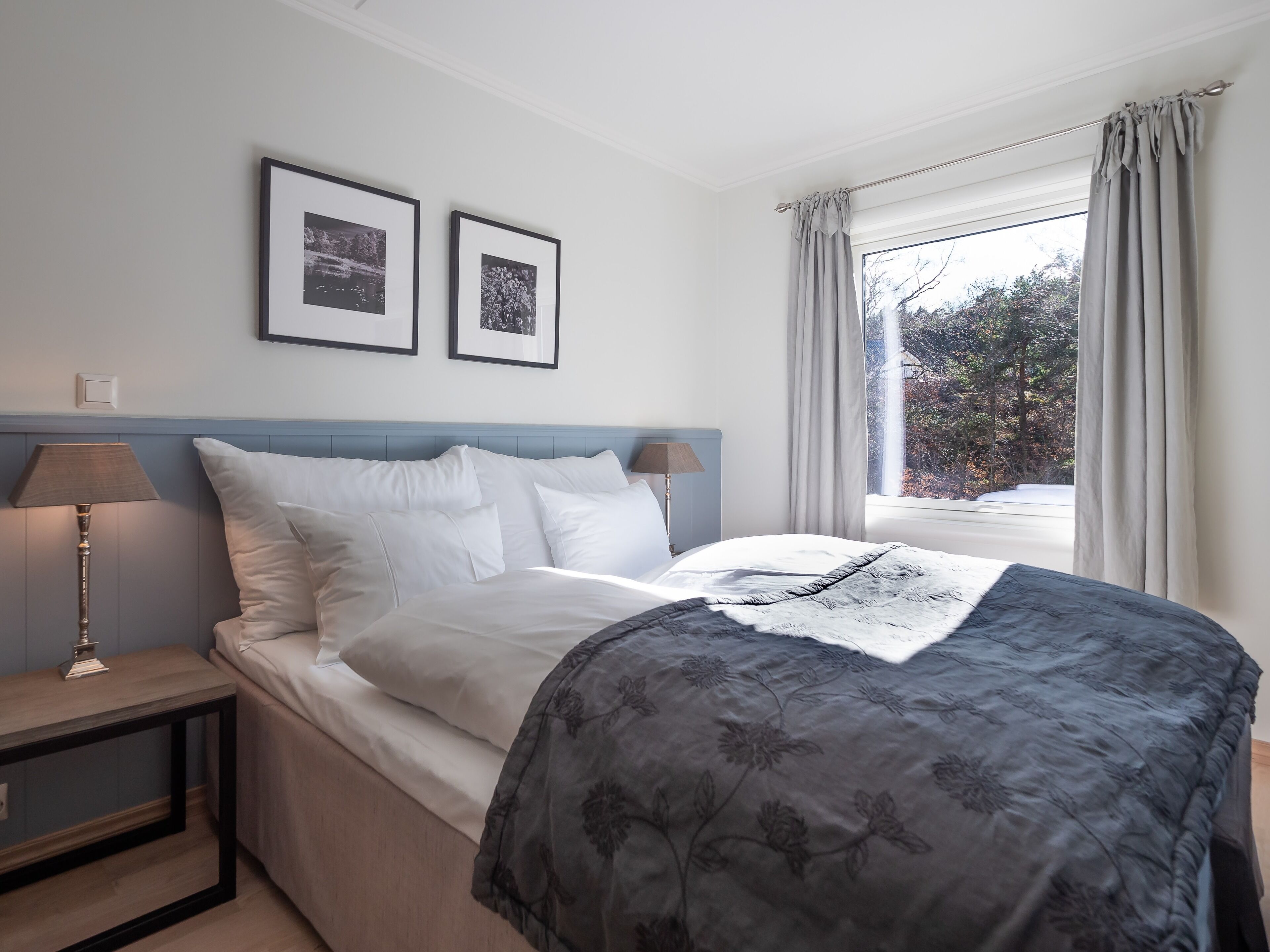 Una cama con ropa de cama gris estampada, dos cuadros en la pared, una lámpara de noche y una ventana con cortinas.