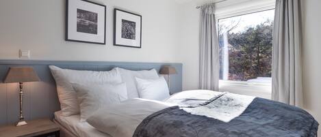 Una cama con ropa de cama gris estampada, dos cuadros en la pared, una lámpara de noche y una ventana con cortinas.