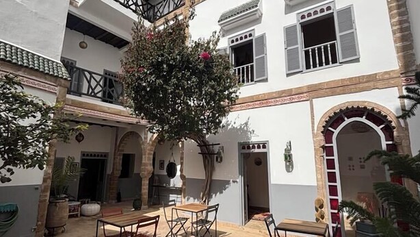 A courtyard with a wooden table and chairs, surrounded by a white building with balconies and windows. - Dar Dayana (Essaouira)
