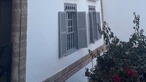 A multi-story building with white walls and grey shutters, featuring a balcony and a small garden area with red flowers. - Dar Dayana (Essaouira)