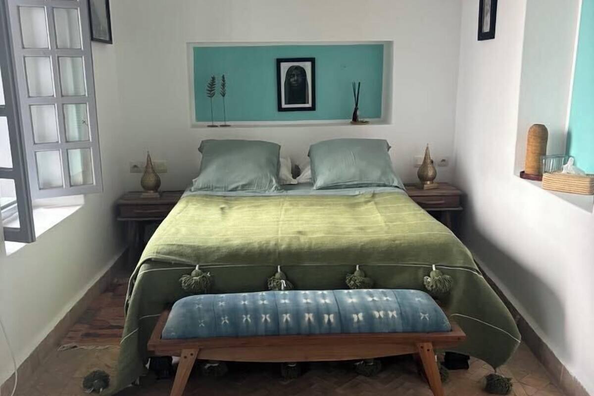 A bedroom with a bed, a bench, a window, and a wall-mounted shelf with decorative items.