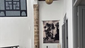 A cozy outdoor seating area with wicker chairs and ottomans, a black metal railing, a hanging pendant light, and a framed black and white photograph on the wall. - Dar Dayana (Essaouira)