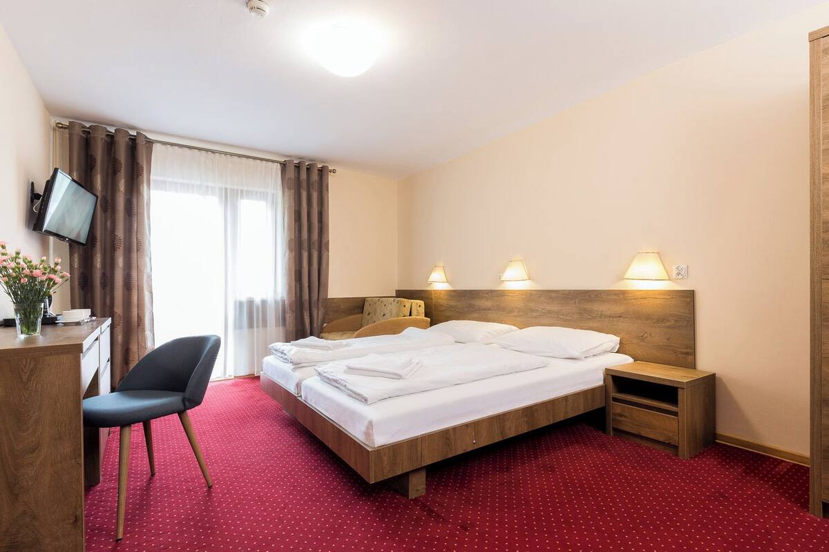 A hotel room with a bed, bedside tables, a desk with a chair, a television, and a window with curtains.