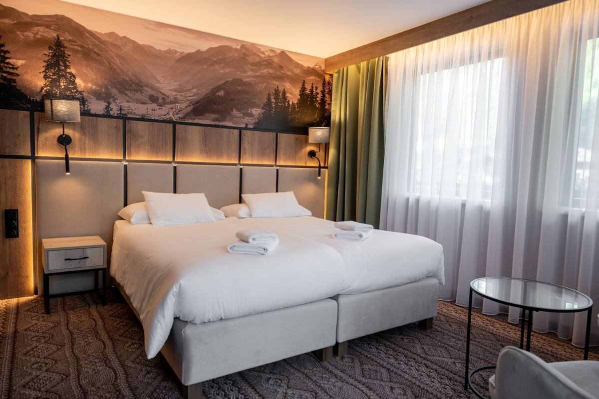 A hotel room with a large bed, a bedside table, a nightstand, a round glass table, and a view of mountains through sheer curtains.
