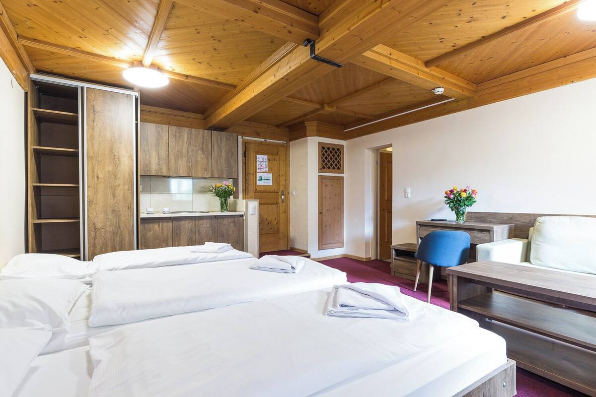 A hotel room with wooden ceilings, a kitchenette, a desk, a chair, and two beds.