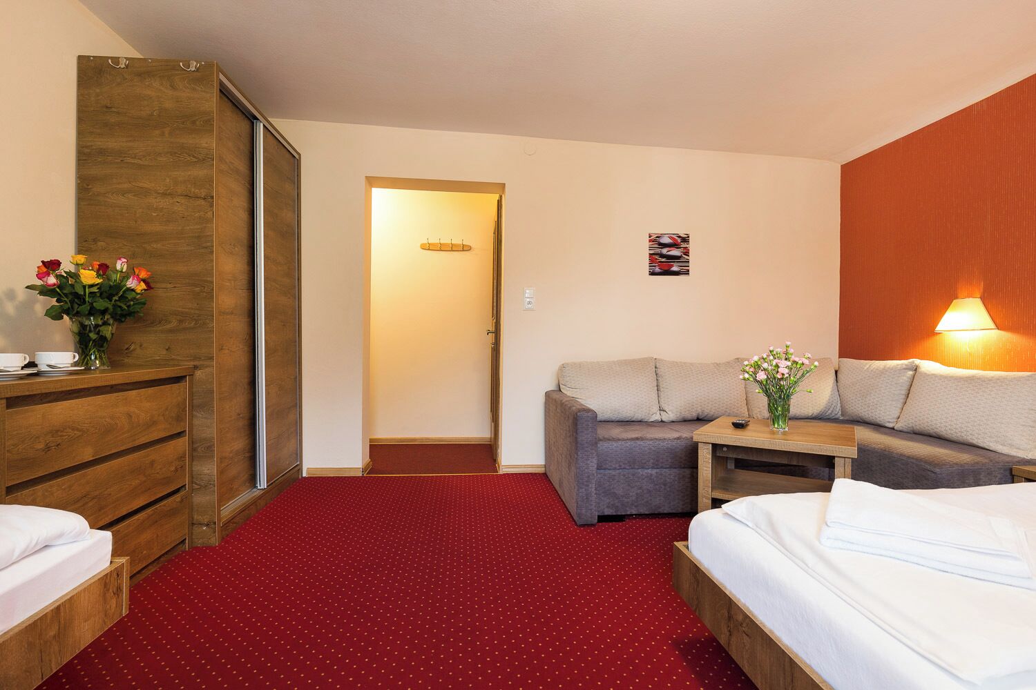 Camera d'albergo con due letti, un settimino in legno, un tappeto rosso, un divano, un tavolino e una lampada fissata al muro.