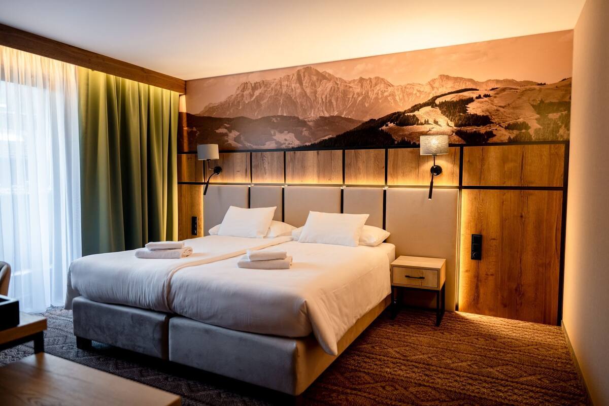 A hotel room with a large bed, two bedside tables, a wooden headboard, and a mountain landscape mural on the wall.