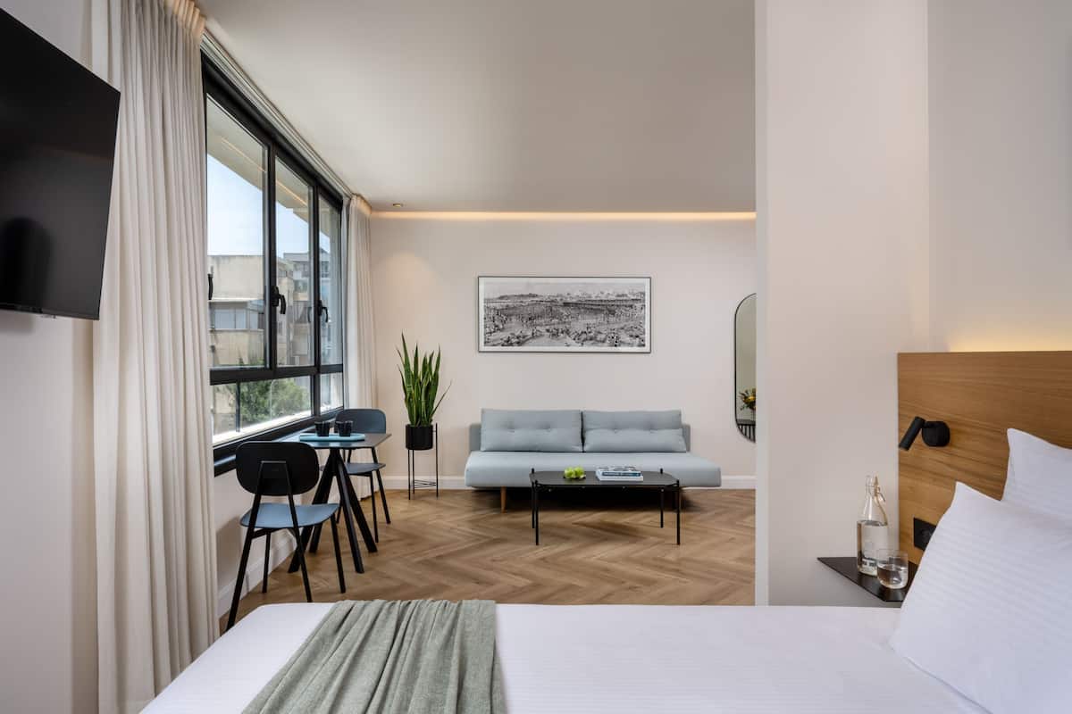 A modern hotel room with a large bed, a sofa, a dining area with a table and chairs, a TV mounted on the wall, and a large window with curtains.