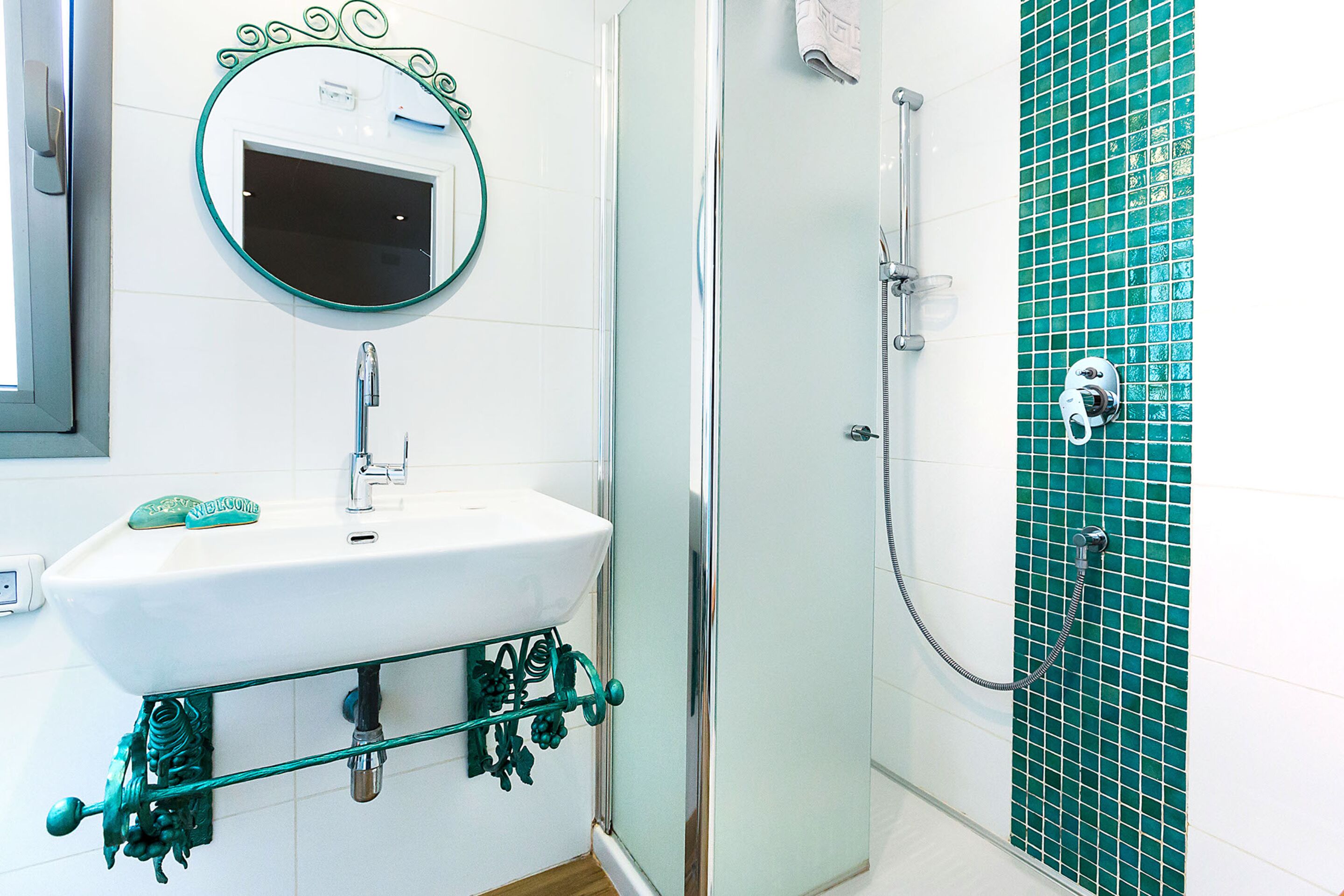 Een badkamer met een wit wastafelmeubel, een ronde spiegel en een douche met een wand betegeld met groene tegels.