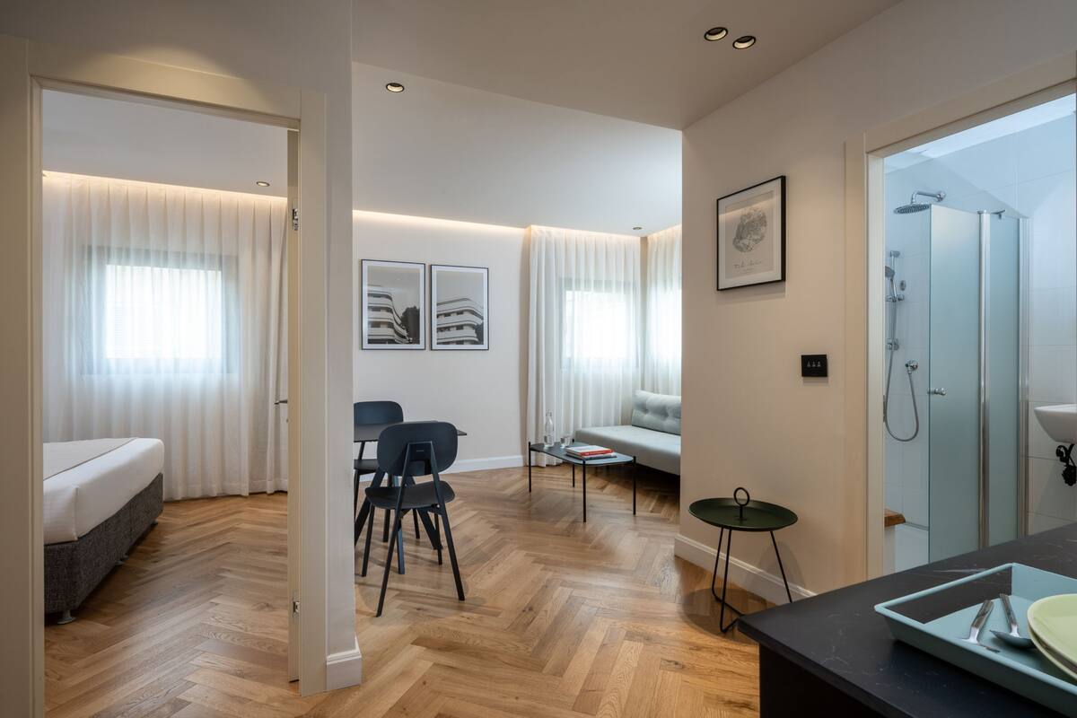 A modern hotel room with a bed, a seating area with a sofa and chairs, a small table, and a shower area.