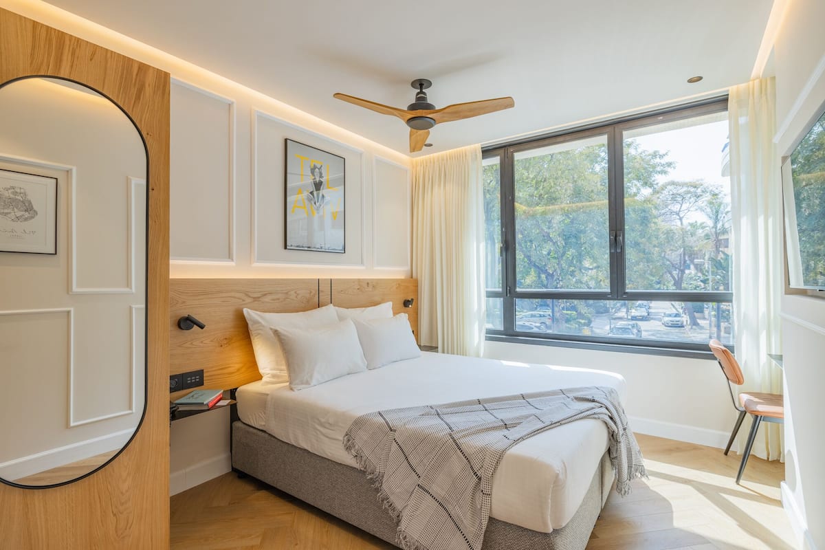 A modern bedroom with a large bed, a ceiling fan, and a window with a view of trees and parked cars.