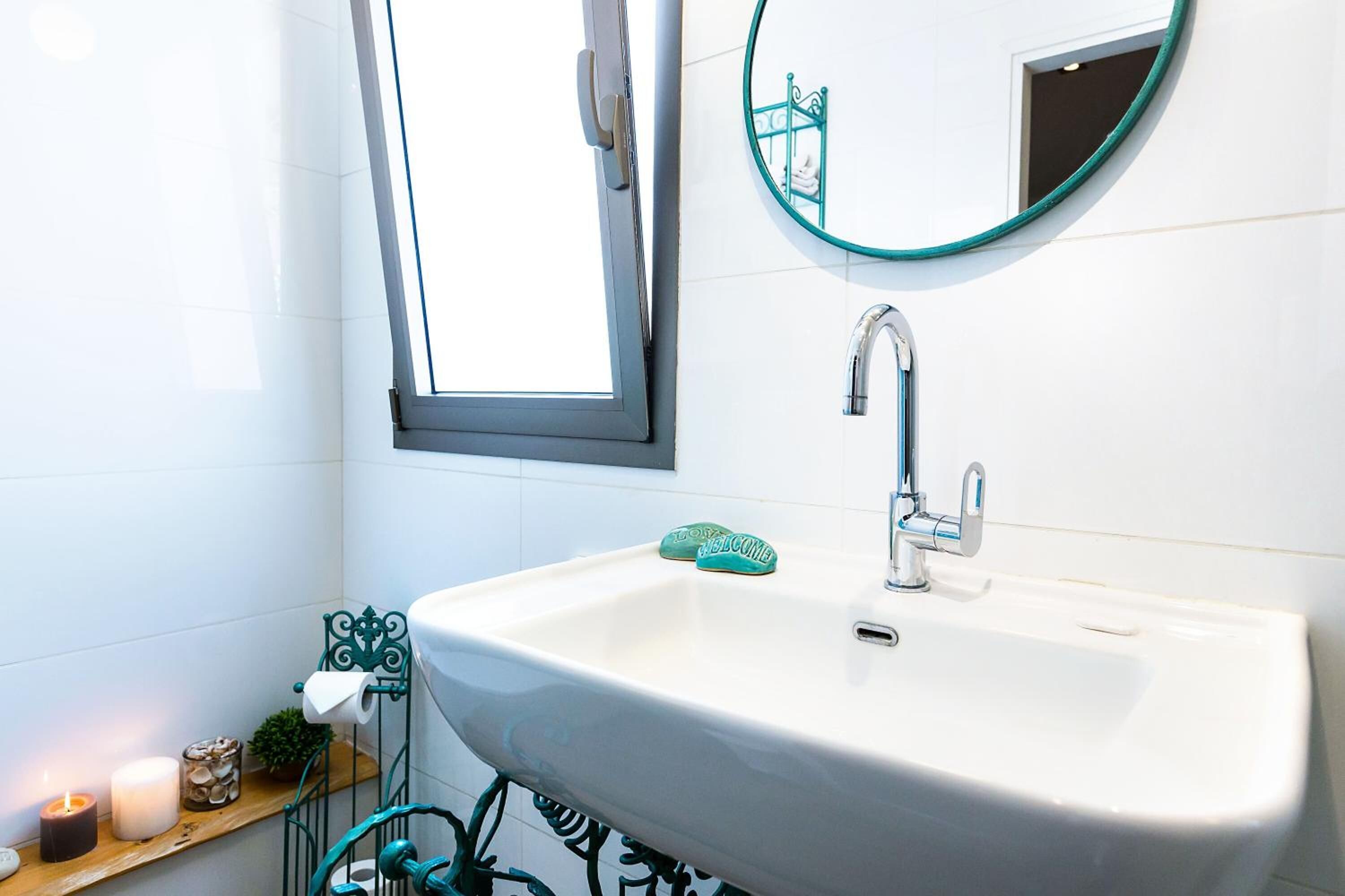 Un baño con lavamanos blanco, ventana, espejo y repisas con artículos de tocador.