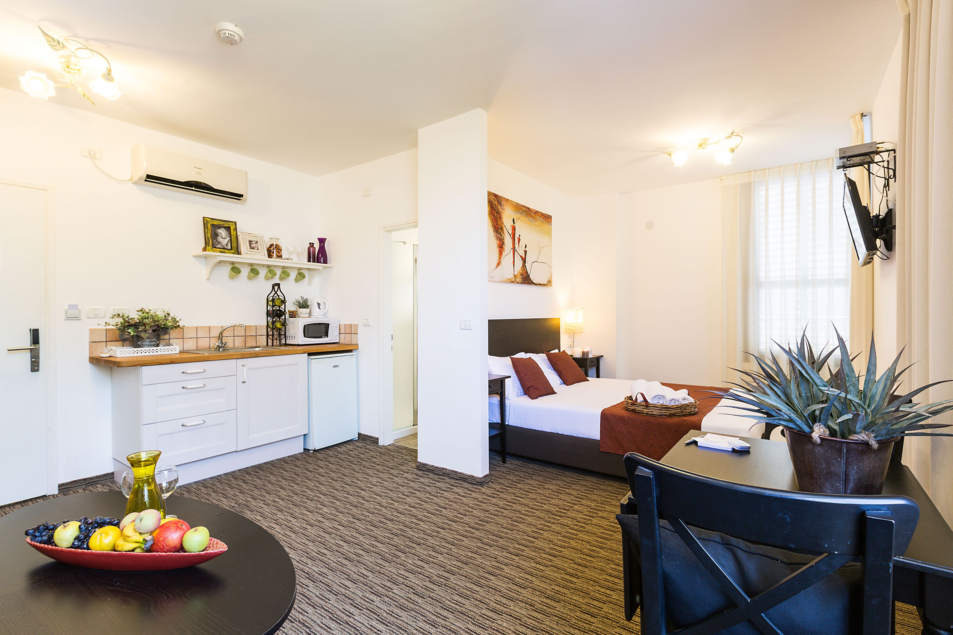 A hotel room with a bed, bedside table, and a dining area with a table and chairs. There is a kitchen area with a microwave and a sink.