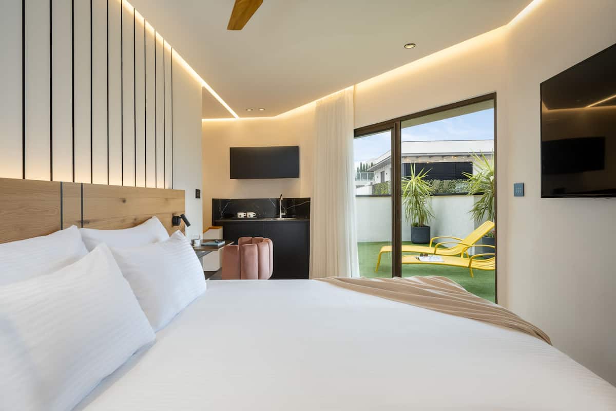 A modern hotel room with a large bed, a flat-screen TV mounted on the wall, a small desk with a chair, and a sliding glass door leading to an outdoor area with a yellow chair.