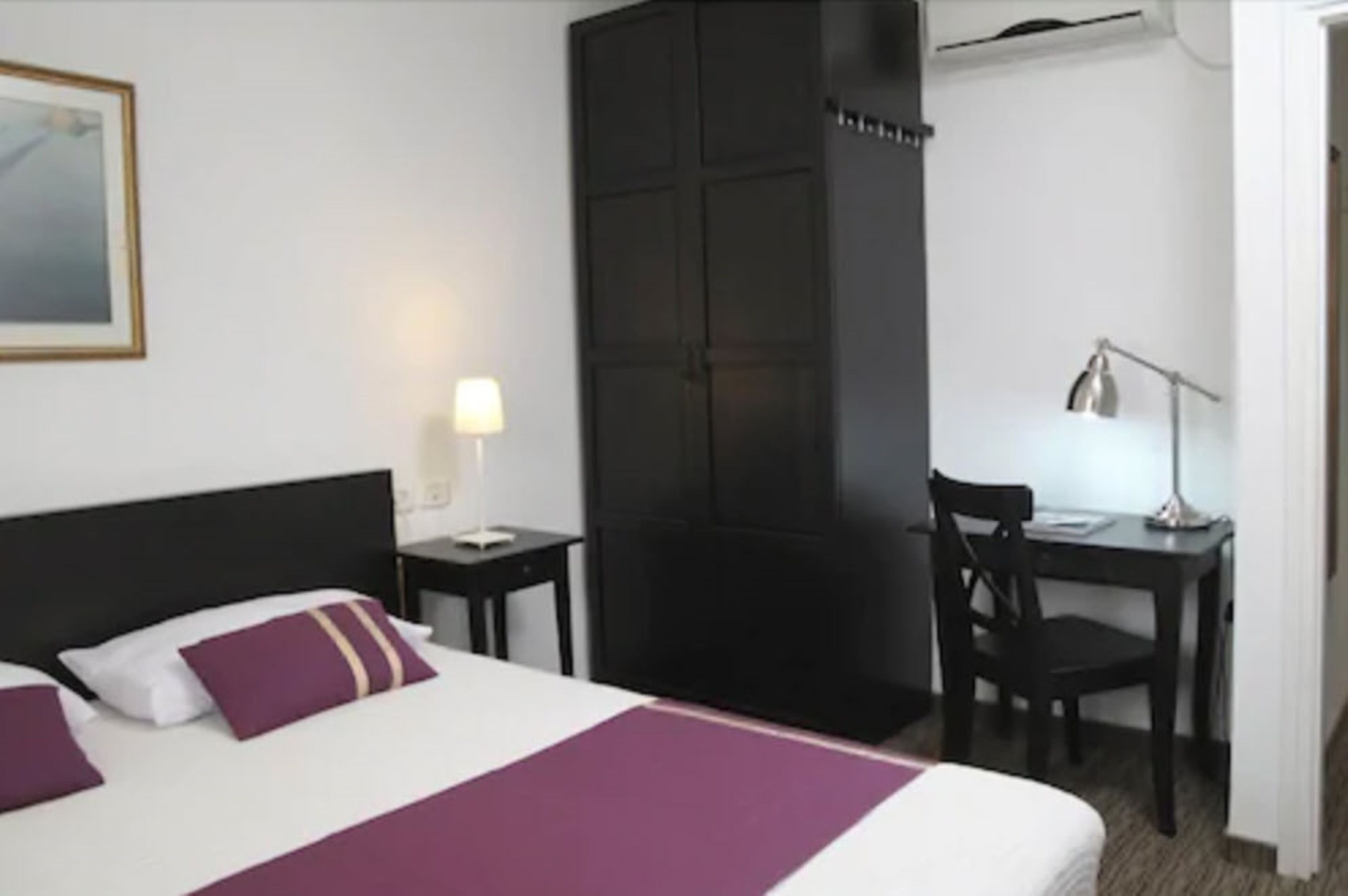 Quarto de hotel com cama, escrivaninha, cadeira, guarda-roupa e abajur.