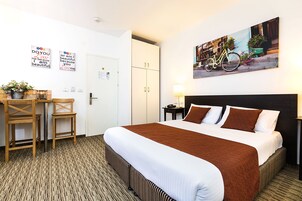 Quarto de hotel com uma cama grande, uma mesa com cadeiras, um vaso de planta e um quadro pendurado na parede.