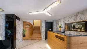 A modern hotel lobby with a wooden reception desk, marble flooring, a staircase, and a wall-mounted mirror.