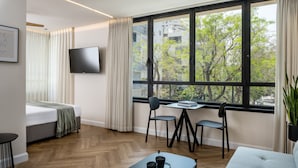 A modern hotel room with a large window, a bed, a desk with two chairs, a TV, and a potted plant.