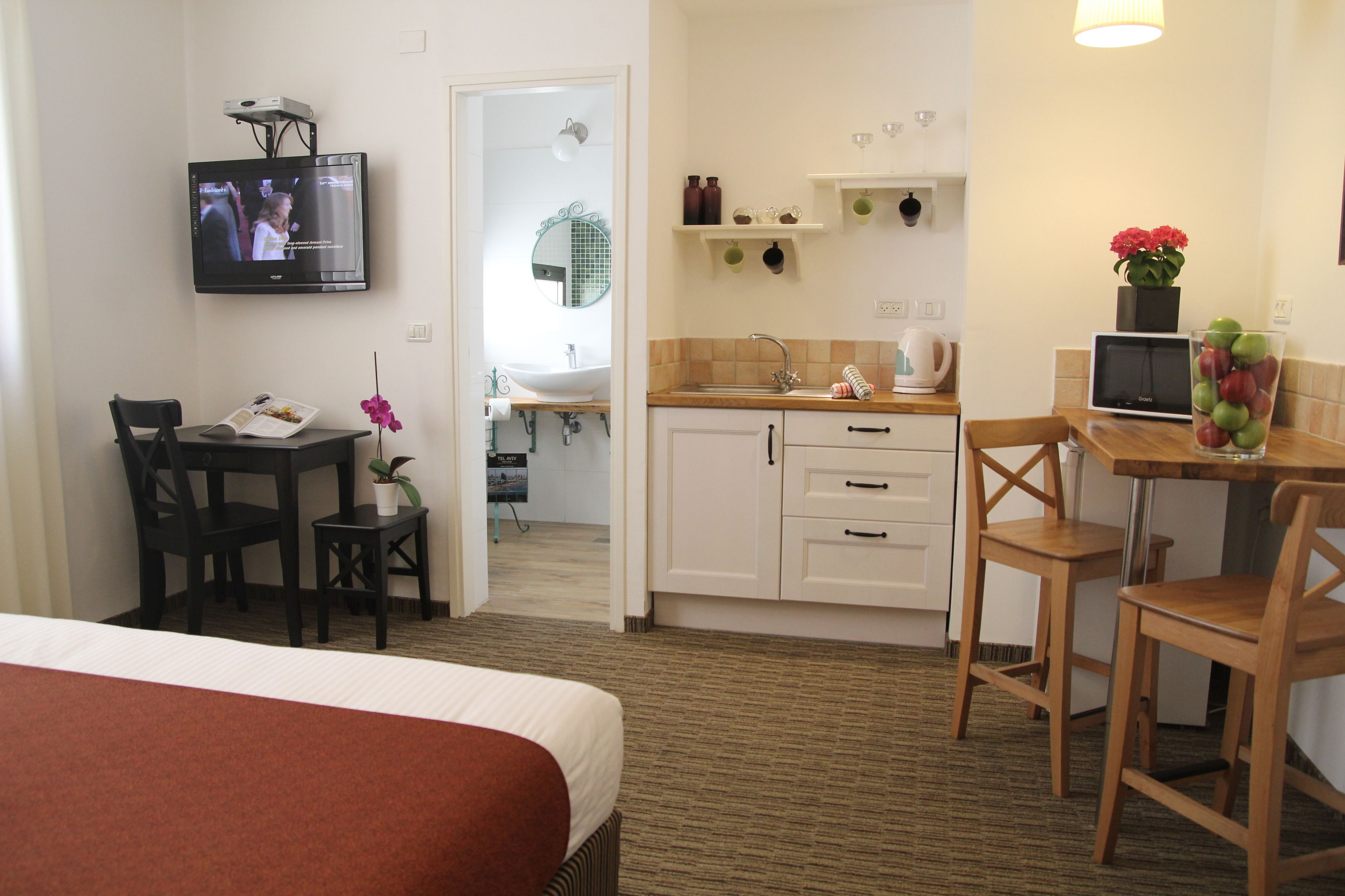 Quarto de hotel com cama, secretária com computador, pequena mesa com cadeiras, banheiro com pia e espelho, e televisão fixada na parede.