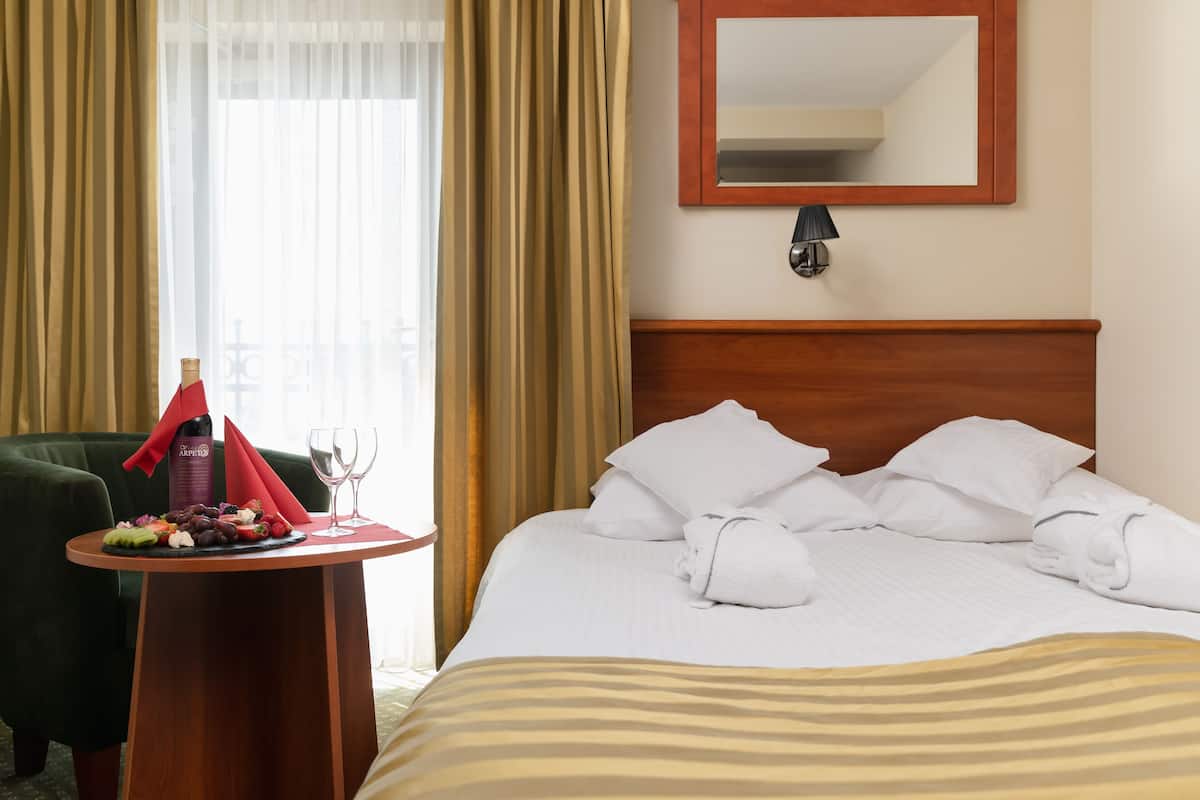 Habitación de hotel con una cama bien hecha, una mesita redonda con una botella de vino, copas y una bandeja de frutas.