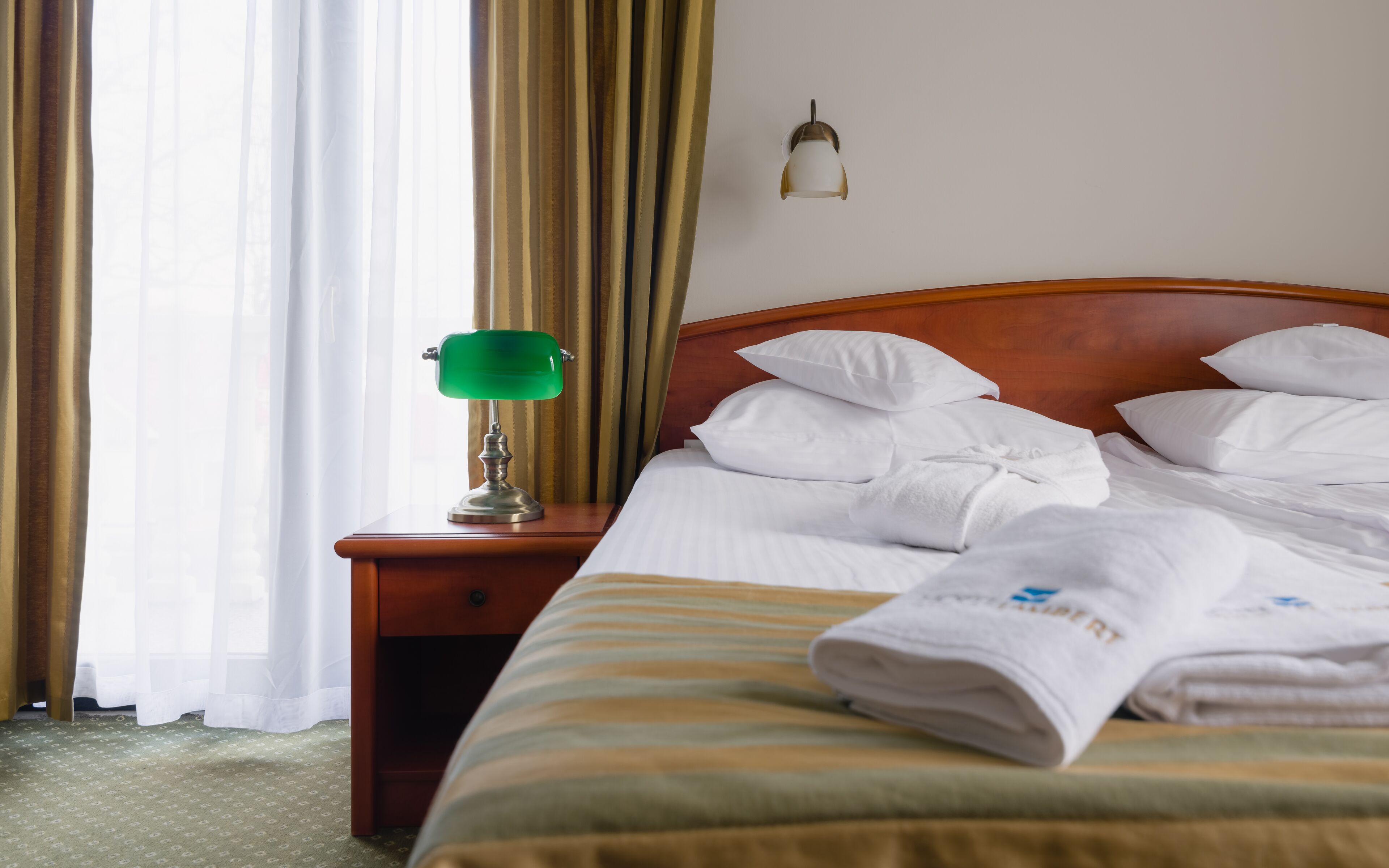 Una cama bien hecha con sábanas blancas y una lámpara de escritorio verde sobre una mesita de noche de madera.