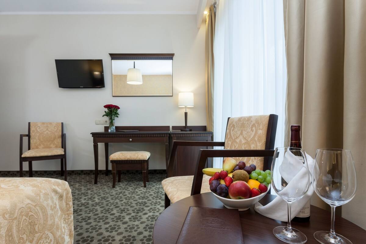 Habitación de hotel con cama, escritorio, silla, televisor, lámpara, ventana con cortinas, y una mesa con frutas y copas de vino.