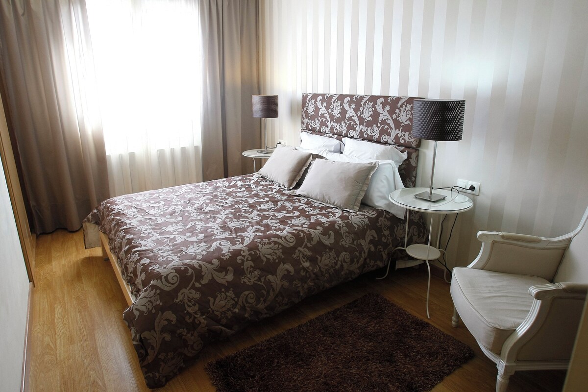 A hotel room with a large bed, two bedside tables, a desk, a chair, and a window with curtains.