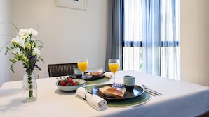 Una mesa de desayuno con un florero de flores, un bol de fruta, dos tostadas, vasos de jugo de naranja y una taza.