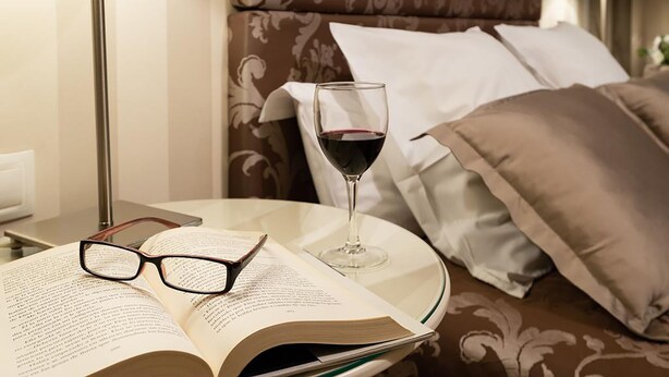 Una cama bien hecha con un cabecero estampado, una mesita de noche con un libro, gafas y una copa de vino tinto.