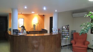 A reception desk with a receptionist, a bookshelf with magazines, and a red armchair. - Tunkelén Apart Hotel (Mendoza)