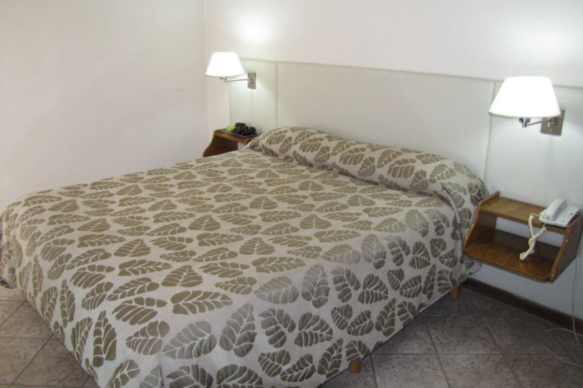 Una cama bien hecha con una colcha estampada con motivos de hojas, dos lámparas de noche y una mesita de noche de madera con un teléfono.