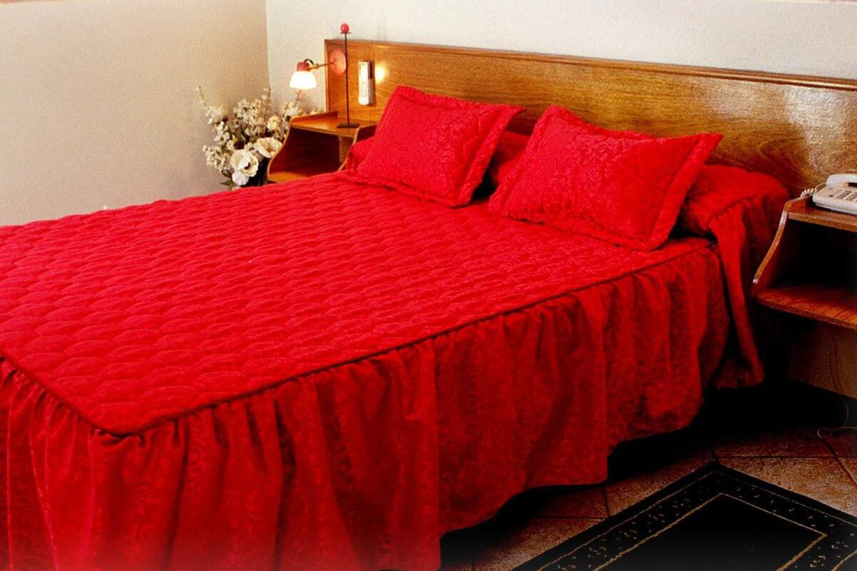 Una cama con colcha y almohadas rojas, cabecera de madera, mesita de noche con lámpara y teléfono.