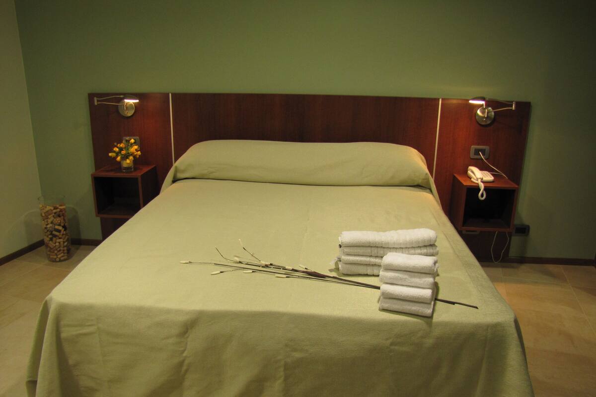 Una cama bien hecha con una colcha verde, una pila de toallas blancas y un jarrón con flores amarillas sobre una mesita de noche.