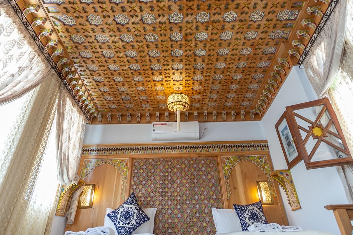 A room with a patterned ceiling, a bed with two pillows, and a wall-mounted air conditioning unit.