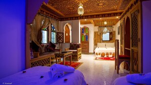 Una habitación tradicional con techo de madera ornamentado, cama con ropa de cama blanca, sofá con cojines estampados y una pequeña mesa con una lámpara.