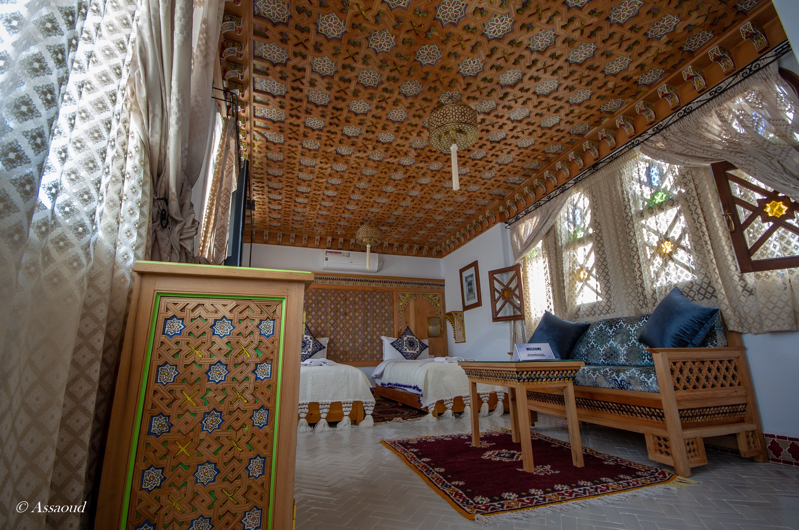 Een kamer met een behangen plafond, twee bedden met behangen dekbedovertrekken, een houten bank met kussens, en een wanddecoratie met een patroon.