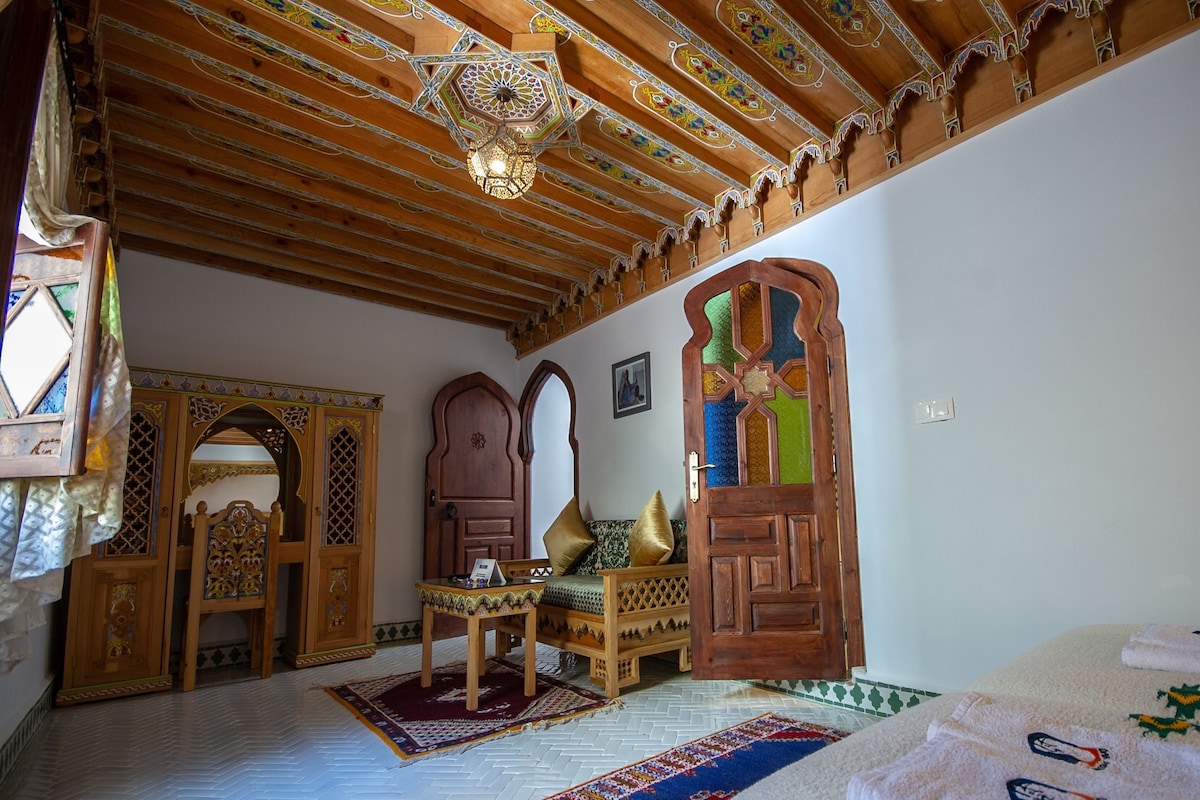 A room with a wooden ceiling, a bed, a table, and a door with intricate designs.