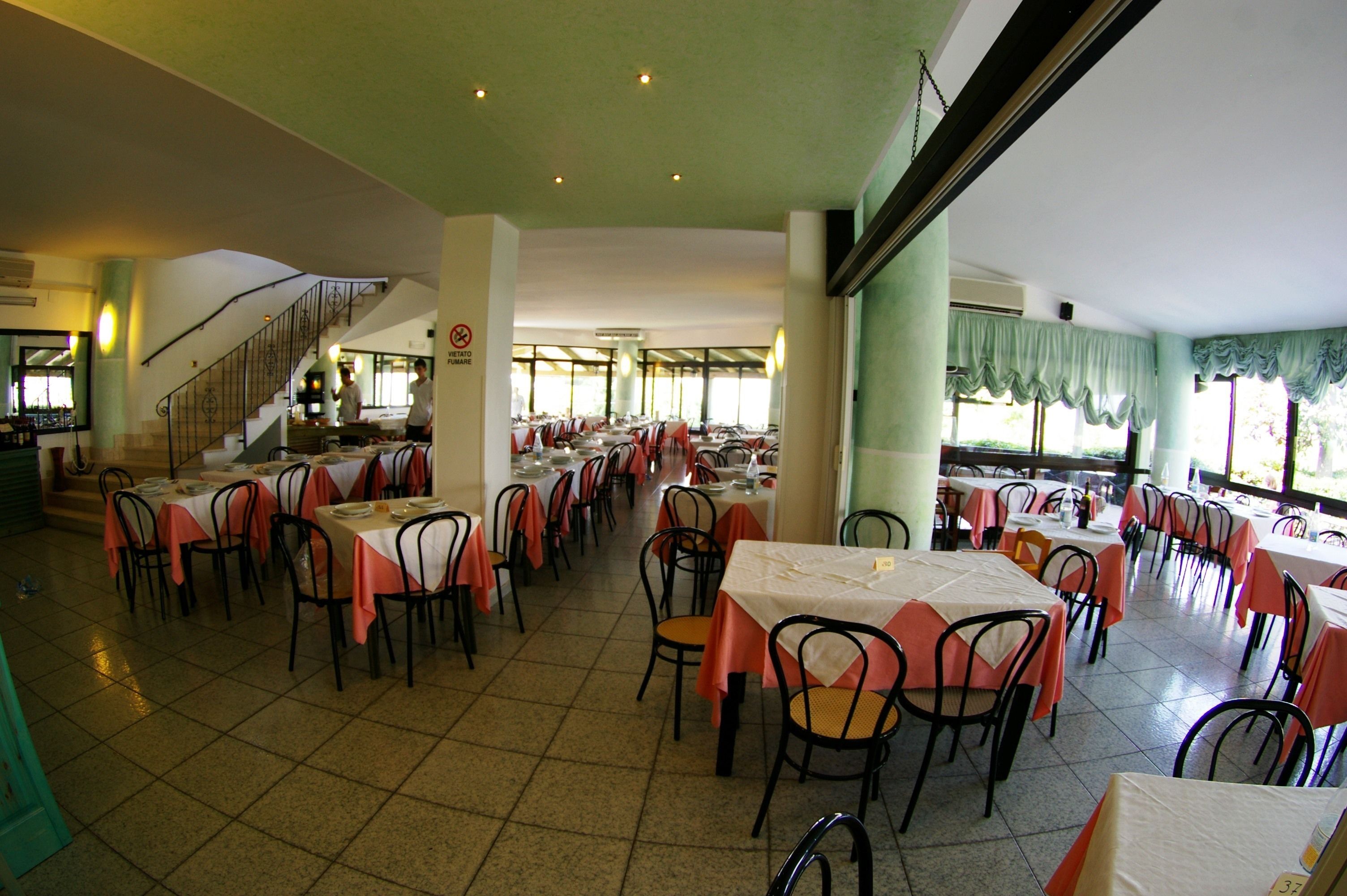 A spacious dining area with multiple tables and chairs, a staircase, and large windows allowing natural light.
