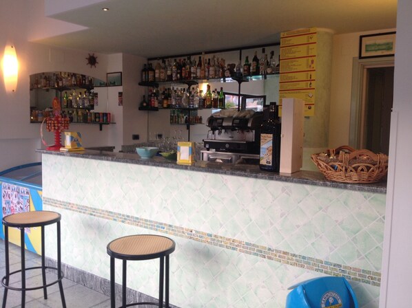A bar area with a counter, shelves of bottles, a coffee machine, and two stools. - Hotel Marinella (Ricadi)