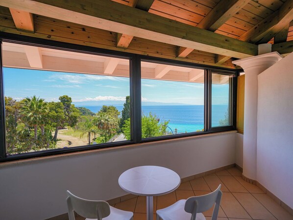 A room with a large window overlooking a coastal landscape, a small round table, and two chairs. - Hotel Marinella (Ricadi)