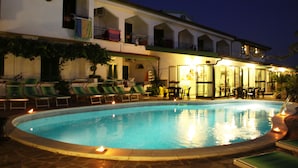 A swimming pool with lounge chairs and tables, surrounded by a building with multiple balconies and a tiled roof.